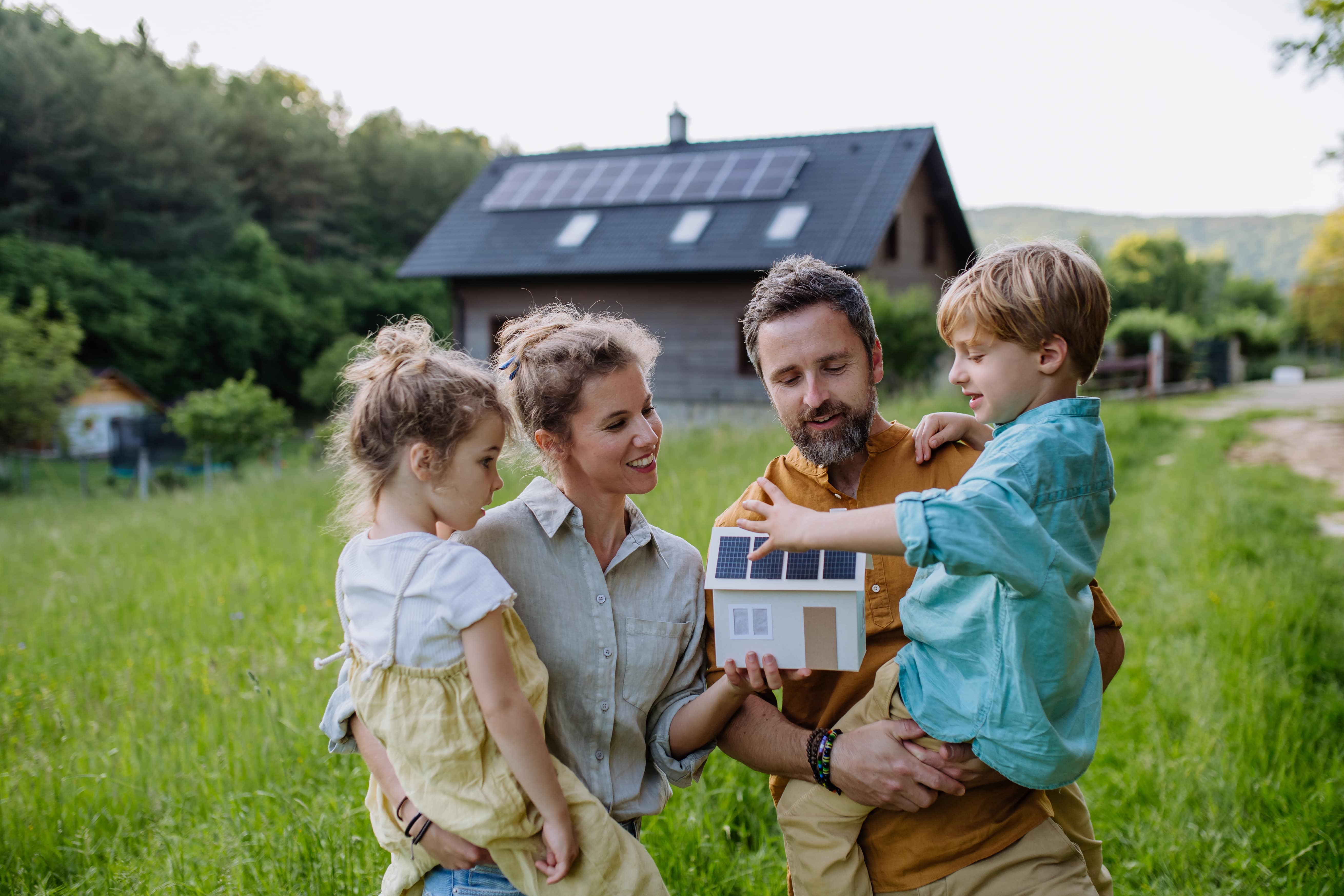 Eine Familie steht im Freien und hält ein Modellhaus mit einer Solaranlage auf dem Dach in der Hand.