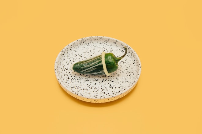 Eine grüne Chilischote mit einem übergezogenen Kondom liegt auf einem gesprenkelten Teller.