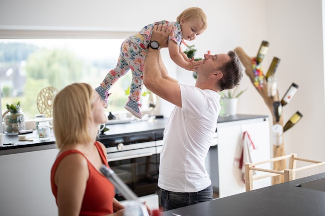 Zwei Eltern in einer modernen Küche, der Vater hebt ein Baby in die Luft