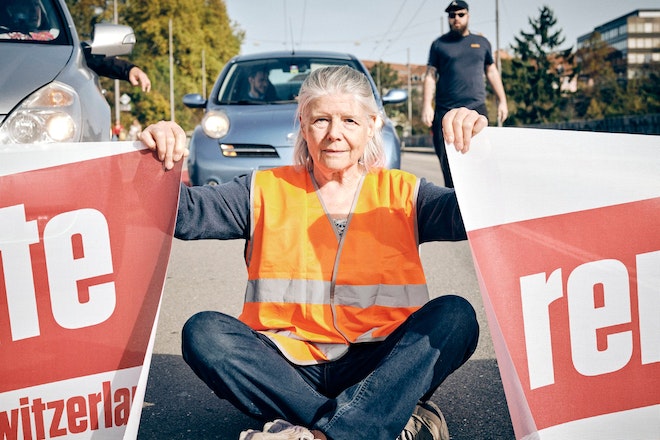 Eine ältere Frau sitzt mitten auf einer Strasse und hält zwei Banner, beschriftet mit dem Begriff 