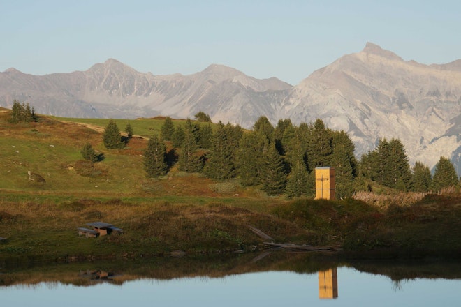 Ein Kompotoi vor einem Bergpanorama spiegelt sich in dem Bergsee.