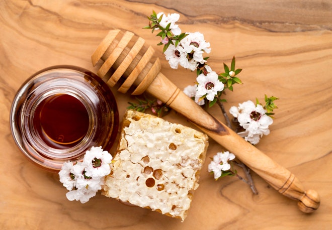 Ein Glas Honig, ein Stück unverarbeiteter Honig, ein paar Blüten der Südseemyrte und einen Honigschöpfer liegen auf einem Holztisch
