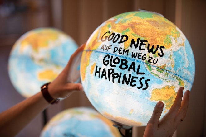 Zwei Hände halten eine leuchtende Weltkugel. Auf der Kugel steht «Good News auf dem Weg zu Global Happiness»