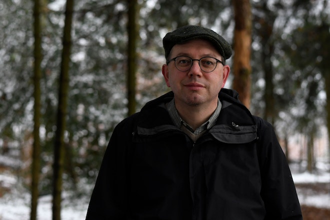 Ein Mann in schwarzer Jacke in Beret und Brille steht in einem verschneiten Wald