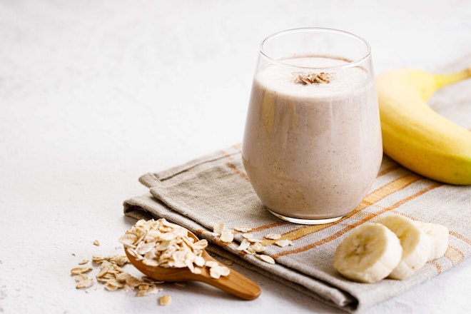 A glass of a light smoothie, next to a banana and oatmeal