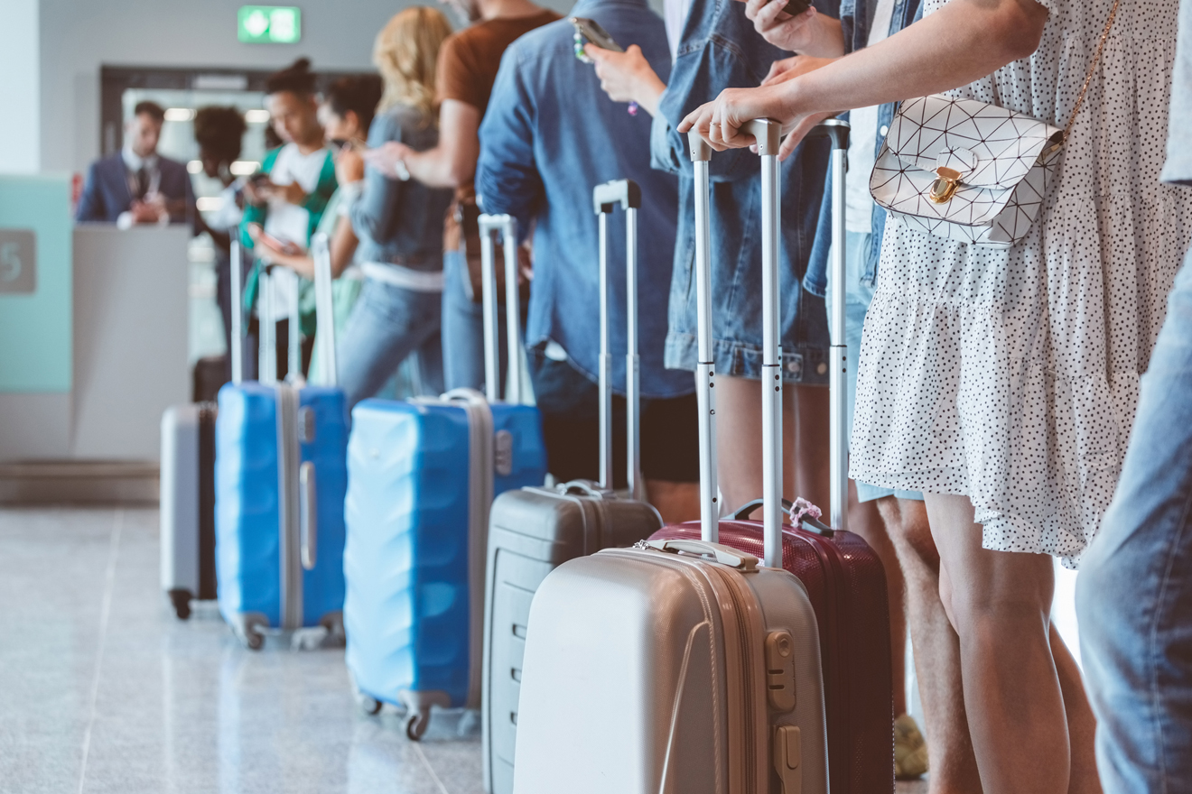 Mehrere Menschen mit Rollkoffer stehen vor der Gepäckabgabe beim Flughafen