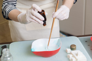 jemand schüttet aus einem Gläschen ein paar Tropfen in eine Schüssel – mit der anderen Hand ein Holzstecken zum Rühren